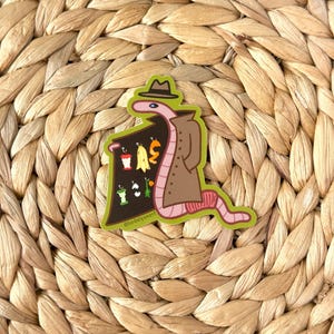 May include: A brown worm wearing a brown trench coat and a brown fedora hat is holding a sign with a variety of fruits and vegetables. The sign is green and has the text "@hadelvyoart" on the bottom.