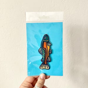 Pu&ograve; includere: Toppa ricamata a forma di pesce blu, arancione e verde, su un cartoncino blu, in una custodia di plastica trasparente. Il pesce ha un design dettagliato e stilizzato.