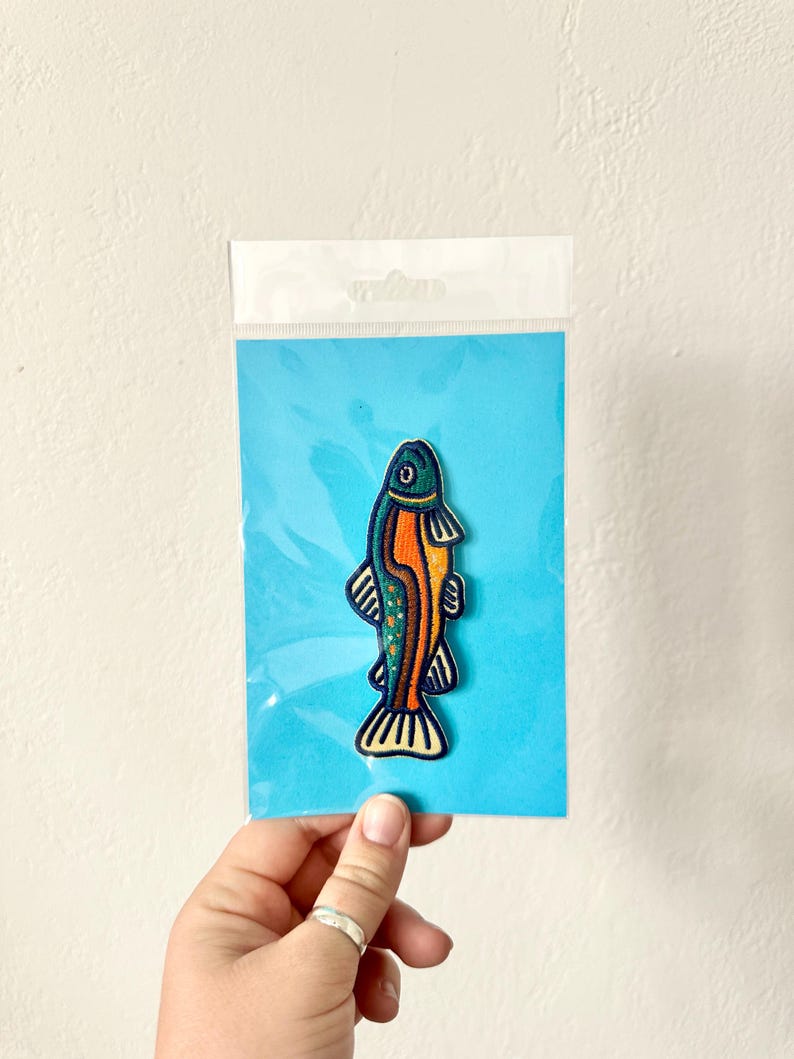Pu&ograve; includere: Toppa ricamata a forma di pesce blu, arancione e verde, su un cartoncino blu, in una custodia di plastica trasparente. Il pesce ha un design dettagliato e stilizzato.