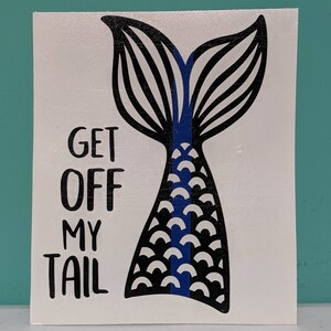 May include: White rectangular sign with a black and blue mermaid tail design. The text "GET OFF MY TAIL" is printed in black, above the tail illustration. The tail has black and white details with a blue stripe.