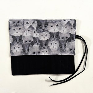 May include: A black pencil roll with a zipper closure and a drawstring. The fabric features a repeating pattern of gray and white cat faces.