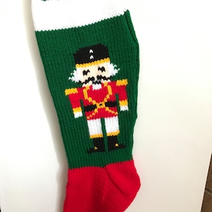May include: A green knitted Christmas stocking with a white cuff and a red toe. A nutcracker design is featured in the centre of the stocking.