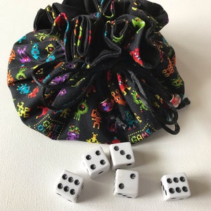 May include: A black fabric dice bag with a colorful pixelated alien pattern and the word "GAME" printed on it. Five white dice are scattered on a white surface.
