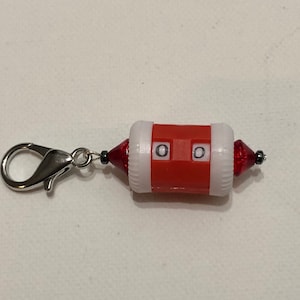 May include: Red and white stitch counter with a silver clip. The counter has two dials with the number "0" on each.