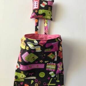 May include: A black fabric hanging organizer with a pink lining and a pink and black fabric pincushion. The organizer has a print of sewing-themed items, including scissors, thread spools, buttons, and a sewing machine. The text "Stitch and Knit" is printed on the organizer.