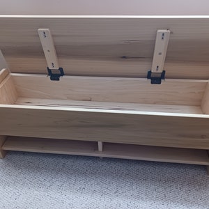 May include: A light-colored wooden storage bench with a hinged lid, open to reveal an empty interior. The bench has a lower shelf and is constructed from unfinished wood. The lid is held open by two black metal hinges.