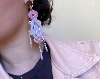 May include: A single earring with a light purple, teardrop-shaped design, accented with pink flowers and gold-colored metal detailing. The earring has a dangling chain with small, white beads and a circular stud.