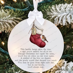May include: A round white Christmas bauble with a red stocking design, a small bird, and the word "Dad" on a tag. The bauble has a white ribbon and bow. Text on the bauble reads: "This bauble hangs upon the tree..."