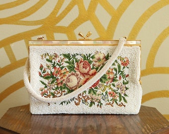 May include: A vintage beaded handbag with a floral tapestry design. The off-white bag features a rectangular shape, a beaded handle, and a gold-toned clasp. The floral design includes pink, red, and green flowers. The bag is displayed on a small wooden table.