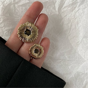 Puede incluir: Dos pendientes de color dorado con un diseño texturizado con forma de flor. Los pendientes tienen forma circular con un círculo abierto más pequeño en el centro.