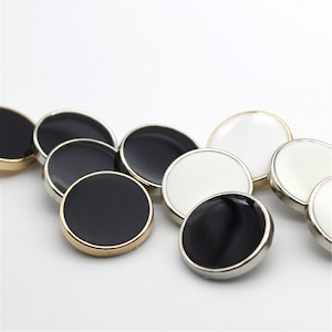May include: A selection of round buttons in black and white with metallic rims. Some buttons have gold-coloured rims, while others have silver-coloured rims. The buttons are arranged on a white background.