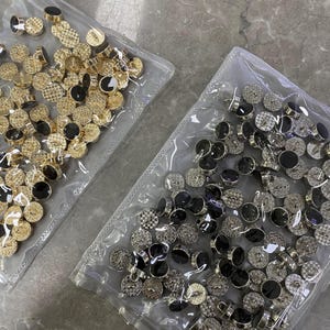 Könnte beinhalten: Zwei Beutel mit Knöpfen, einer mit goldenen und schwarzen Knöpfen, der andere mit silbernen und schwarzen Knöpfen.