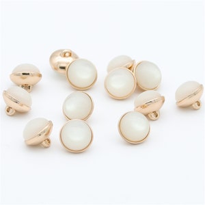 Puede incluir: Un conjunto de diez botones blancos perlados con bordes de metal dorado. Los botones tienen una parte posterior plana y un pequeño agujero para coser.