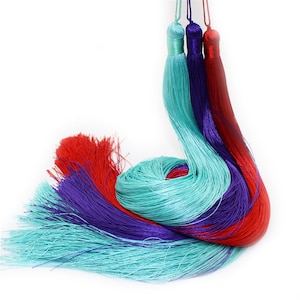 Op de afbeelding: Drie lange kwastjes in turquoise, paars en rood. De kwastjes zijn gemaakt van dunne draden stof en zijn aan de bovenkant aan elkaar gebonden.