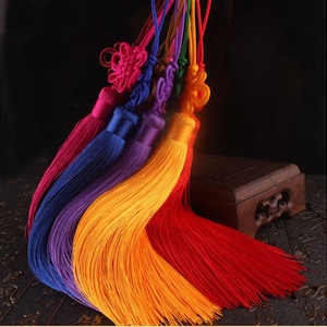 Könnte beinhalten: Ein Set aus sechs bunten Quasten-Schlüsselanhängern mit komplizierten Knotendesigns. Die Quasten bestehen aus seidenen Fäden in Rot, Orange, Gelb, Lila, Blau und Pink.