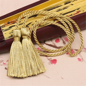 Peut inclure: Deux glands dorés avec un cordon en corde dorée. Les glands sont faits de fils doux et soyeux et sont attachés à une corde tressée.