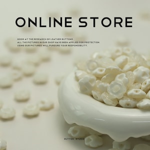 May include: A white ceramic bowl filled with white flower-shaped buttons. The bowl is sitting on a white surface. The text "ONLINE STORE" and "BUTTON STUDIO" is visible in the image.