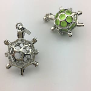 Puede incluir: Dos colgantes de jaula de tortuga plateados. Un colgante está vacío y el otro tiene un orbe de vidrio verde en su interior.