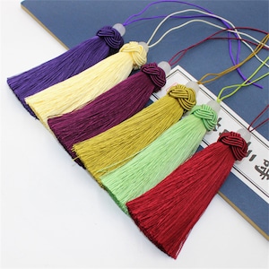 Puede incluir: Seis adornos de borla de colores con diferentes hilos de colores, incluyendo morado, amarillo, burdeos, verde y rojo. Cada borla tiene una pequeña cuenta blanca en la parte superior.