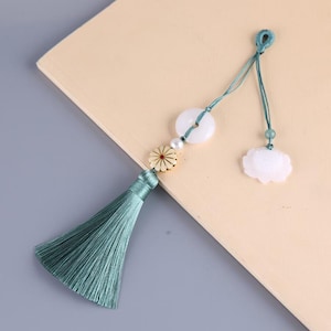 Pode incluir: Um pendão verde-azulado com uma corda decorativa com um pendente circular branco, uma pequena flor, uma pérola e um berloque em forma de nuvem. O pendão é feito de fios sedosos e provavelmente é usado como elemento decorativo.