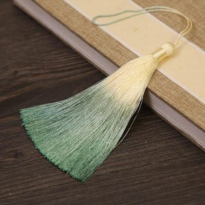May include: A decorative tassel with a gradient of colors, transitioning from pale yellow to green. The tassel is attached to a thin cord and rests on a book with a beige cover and a cream-colored stripe.
