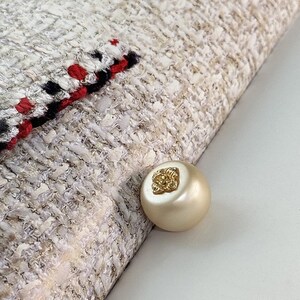 Könnte beinhalten: Nahaufnahme eines cremefarbenen Tweedstoffs mit einer dekorativen Bordüre aus Rot, Schwarz und Weiß. Ein großer, runder, perlenartiger Knopf mit goldenem Blumenmuster ist zu sehen.