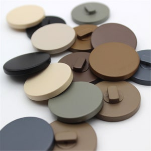 May include: Assortment of round, flat, decorative buttons in various colors, including black, brown, beige, gray, and blue.