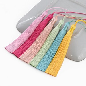 Könnte beinhalten: Sechs bunte Seidenquasten mit langem Fransen. Die Quasten sind in einer Reihe angeordnet, wobei die hellste Farbe oben und die dunkelste Farbe unten ist. Die Farben sind rosa, hellrosa, hellgrün, hellblau und gelb.
