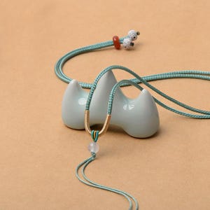 Può includere: Una delicata collana con un cordino azzurro e un accento dorato. La collana presenta un pendente rotondo e trasparente ed è esposta su un supporto in ceramica azzurra. Il cordino è lungo circa 50 cm.