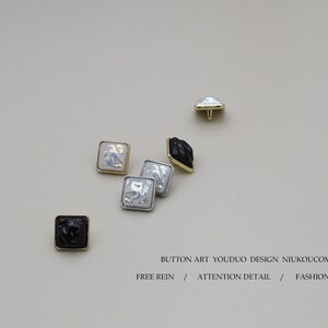 May include: Assortment of square-shaped buttons with a textured, raised design. The buttons are white, black, and silver with gold accents. The buttons are arranged on a white background.