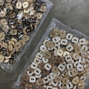 Könnte beinhalten: Ein Nahaufnahmebild von zwei Beuteln, die mit goldenen und schwarzen Knöpfen gefüllt sind. Die Knöpfe sind quadratisch mit einem erhöhten Rand und einer goldenen Oberfläche. Einige der Knöpfe haben eine schwarze Mitte.