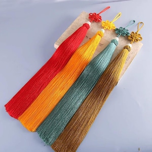 Puede incluir: Una colección de borlas de seda coloridas en rojo, naranja, verde azulado y dorado. Cada borla está rematada con un nudo decorativo y una pequeña cuenta blanca. Las borlas están dispuestas sobre una superficie marrón claro.