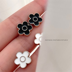 Puede incluir: Un par de pendientes de tono dorado con diseños de flores de esmalte negro y blanco. Las flores tienen un centro circular simple y seis pétalos.
