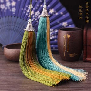 May include: Two decorative tassels with silver-coloured metal caps and colourful threads. The tassels are green, yellow, and teal. The background includes a blue fan and a brown ceramic cup.