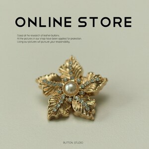 Puede incluir: Botón dorado en forma de flor con un centro de perla y diamantes de imitación. El botón está sobre un fondo beige claro.