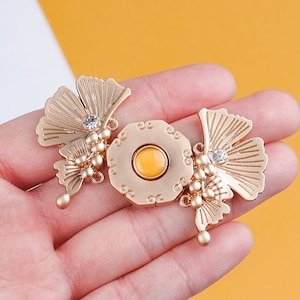 May include: Gold-tone metal brooch with a yellow stone in the center, surrounded by a decorative design. The brooch is shaped like two ginkgo leaves with a beaded detail.