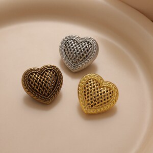 May include: Three heart-shaped decorative objects in gold, silver, and bronze. Each heart features a woven basket design in the center and a braided border. The objects are arranged on a light pink surface.