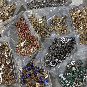 Könnte beinhalten: Sortiment dekorativer Knöpfe in transparenten Plastiktüten. Die Knöpfe haben ein kreisförmiges Design mit einem Sternmuster in verschiedenen Farben, darunter Gold, Rot, Schwarz, Blau und Grün. Die Knöpfe scheinen aus Metall zu sein.