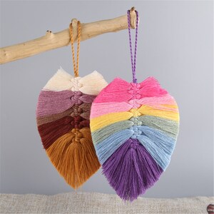 May include: Two colorful macrame leaf wall hangings with fringe. One is a rainbow color scheme and the other is a mix of brown, purple, and cream.