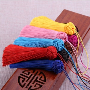 Peut inclure: Assortiment de glands colorés avec des extrémités nouées. Les glands sont faits de fils doux et soyeux dans des tons de jaune, rose, bleu, rouge et bleu marine.