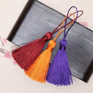 Puede incluir: Tres adornos de borla en color burdeos, naranja y morado. Cada borla tiene una cuerda larga.