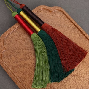 May include: Three tassel ornaments with different colored threads. The tassels are green, dark green, and brown. The tassels are on a wooden surface.