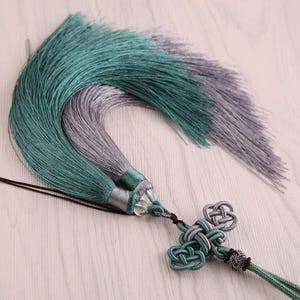 May include: Two decorative tassels in teal and grey. The tassels are made of silky threads and are attached to a decorative knot with a clear bead. These tassels are likely used for home decor or craft projects.
