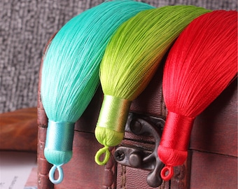 Puede incluir: Tres adornos de borla coloridos con hebras largas y sedosas. Las borlas son turquesa, verde y roja. Se muestran en una caja de madera con un cierre de metal.