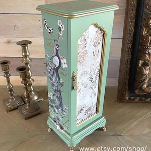May include: A mint green cabinet with gold trim and a peacock design. The cabinet has a patterned door and ornate gold hardware. The top of the cabinet has gold lettering. The cabinet is on small gold feet.