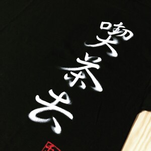 May include: Black t-shirt with white Japanese calligraphy. The design features bold, stylized characters and a red square stamp at the bottom. The shirt is made of a soft material.