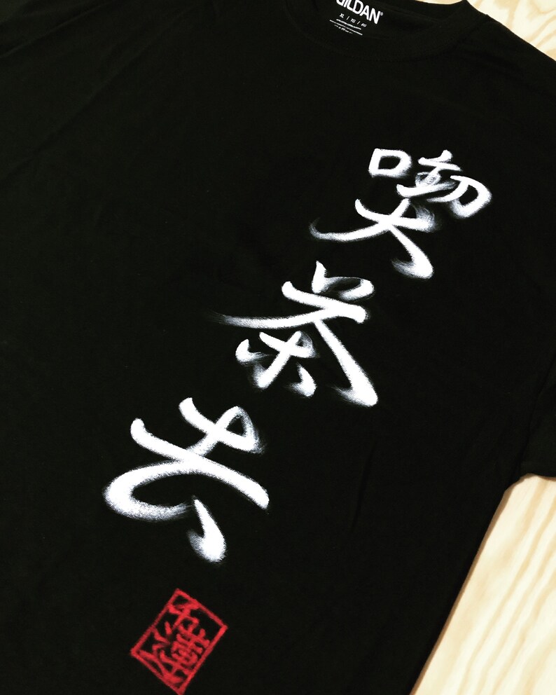 May include: Black t-shirt with white Japanese calligraphy. The design features bold, stylized characters and a red square stamp at the bottom. The shirt is made of a soft material.