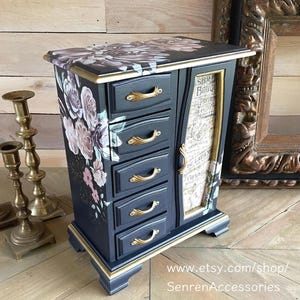 May include: A dark charcoal jewellery armoire featuring five drawers with gold-toned handles, a side cabinet door, and a floral decal on the top and side. The cabinet door displays vintage-style French typography, and the base is accented with gold trim.