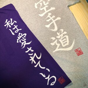 May include: Two t-shirts, one gray and one purple, with Japanese characters printed in white. The gray shirt has "Karate-Do" written on it, and the purple shirt has text on the sleeve. Both shirts have a red stamp.