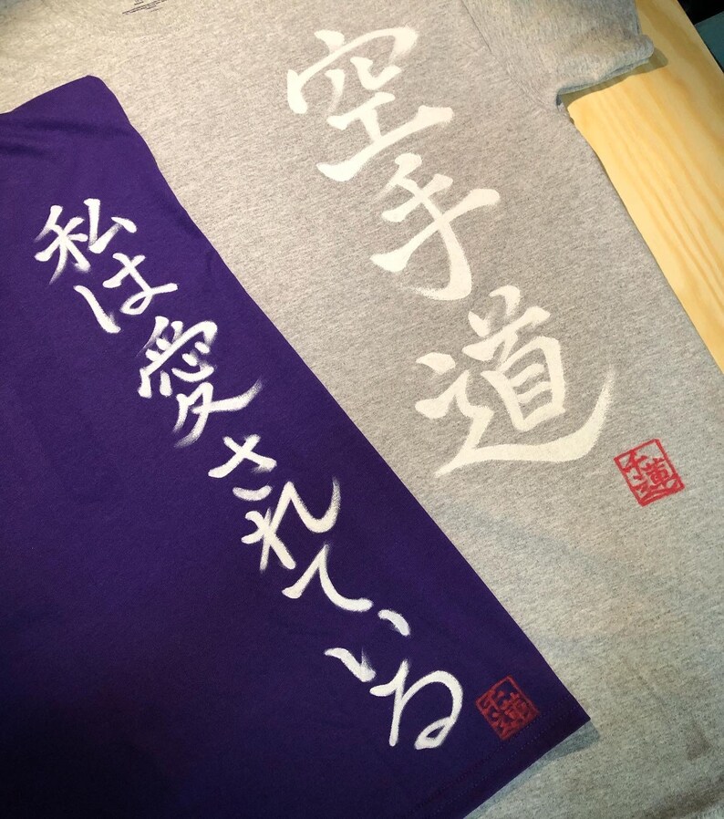 May include: Two t-shirts, one gray and one purple, with Japanese characters printed in white. The gray shirt has "Karate-Do" written on it, and the purple shirt has text on the sleeve. Both shirts have a red stamp.